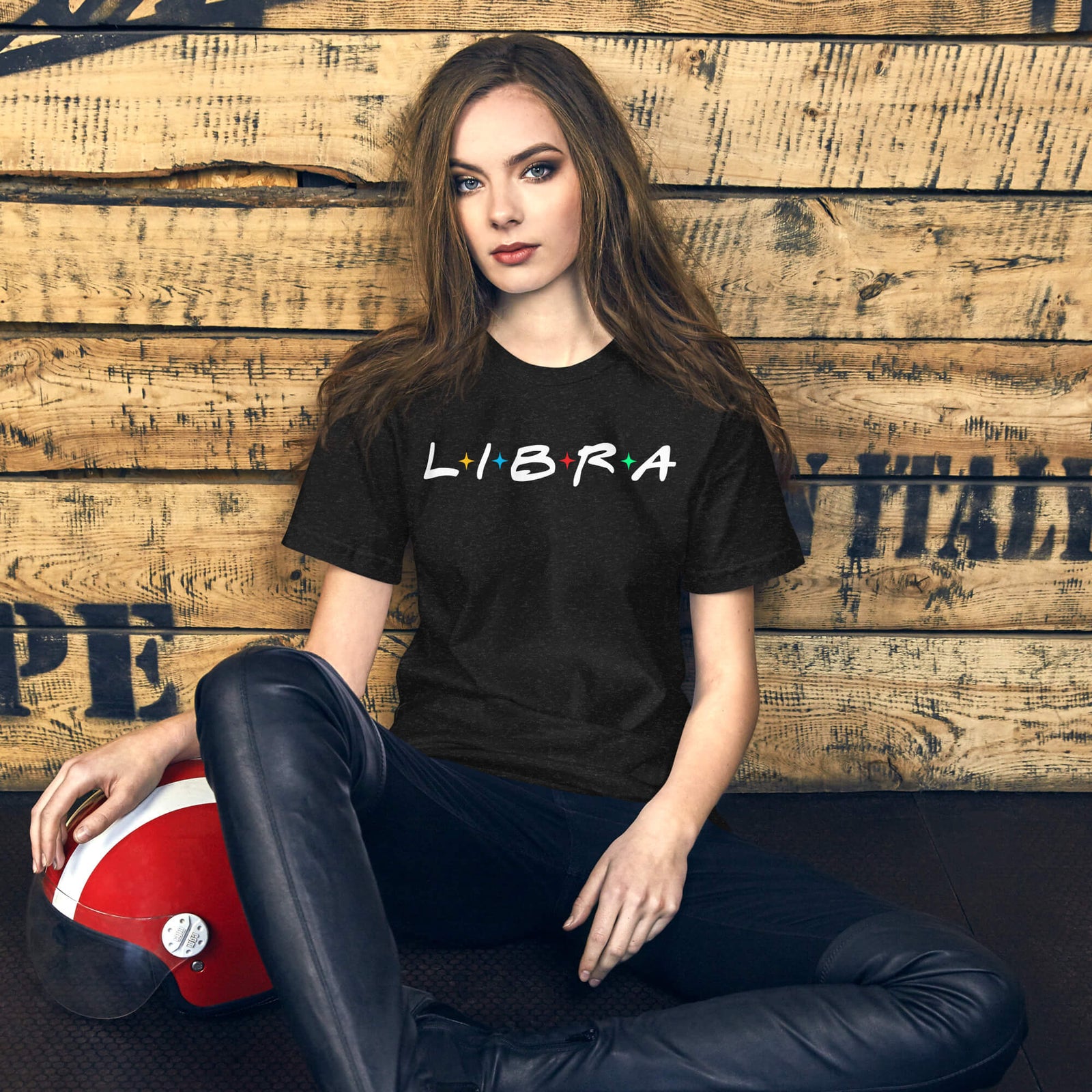 Woman sitting against a wooden wall wearing a black “LIBRA” t-shirt with multicolored stars, next to a red helmet.