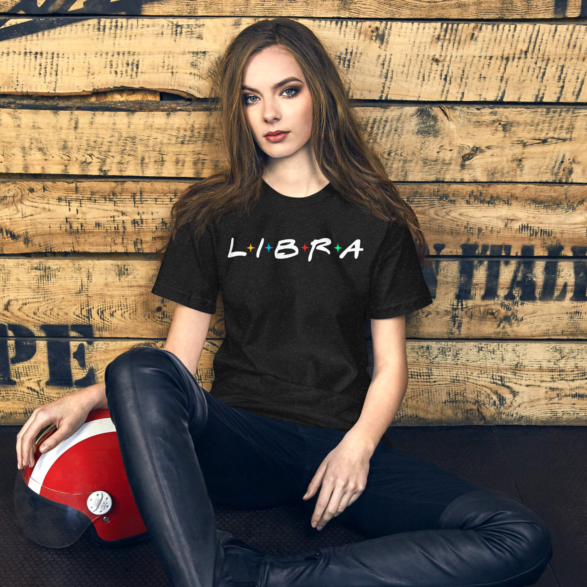 Woman sitting against a wooden wall wearing a black “LIBRA” t-shirt with multicolored stars, next to a red helmet.