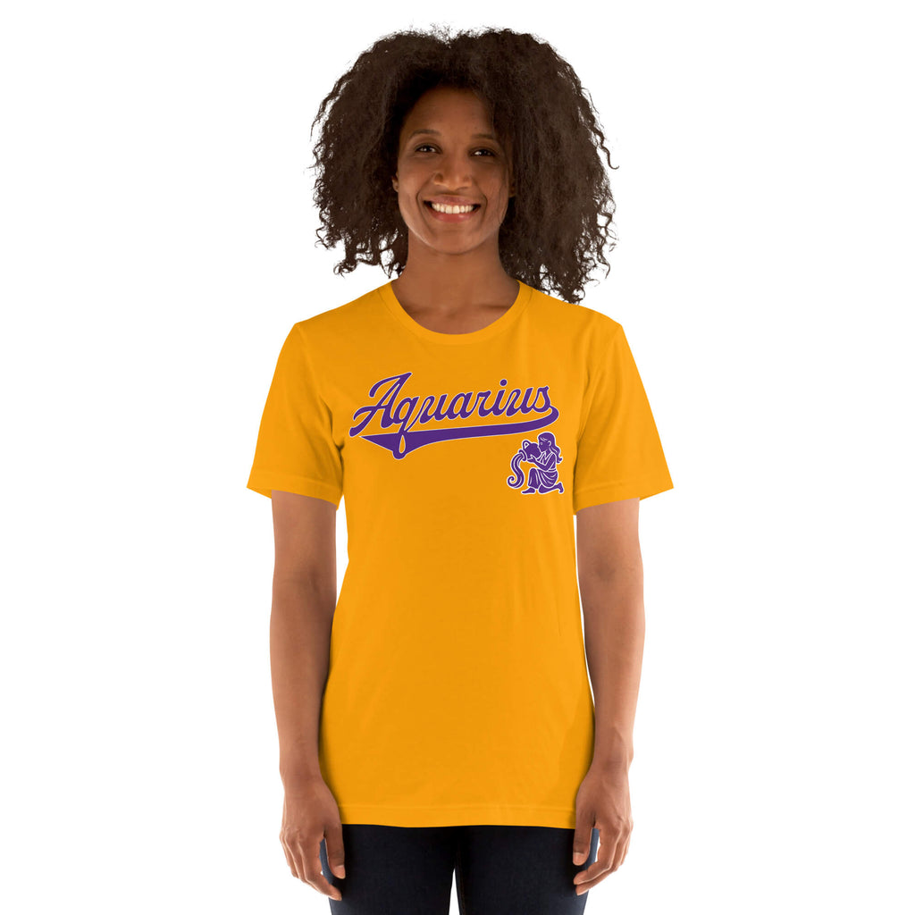 A smiling woman with curly dark hair wearing a gold short-sleeve t-shirt with a purple 'Aquarius' script and zodiac graphic on the chest, set against a plain white background.
