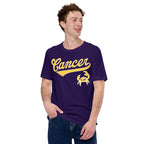 A man wearing a dark purple t-shirt with a gold 'Cancer' script and crab graphic, paired with blue jeans, set against a plain white background.