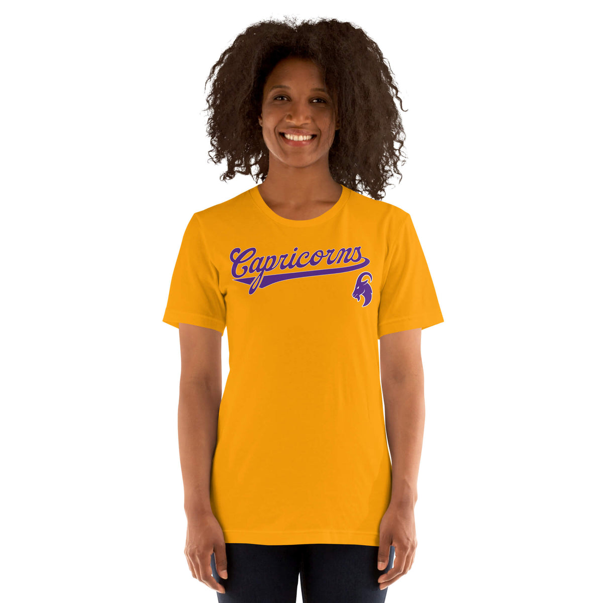 A smiling woman with curly dark hair wearing a bright gold t-shirt with 'Capricorns' written in purple script across the chest, paired with dark pants, set against a plain white background.
