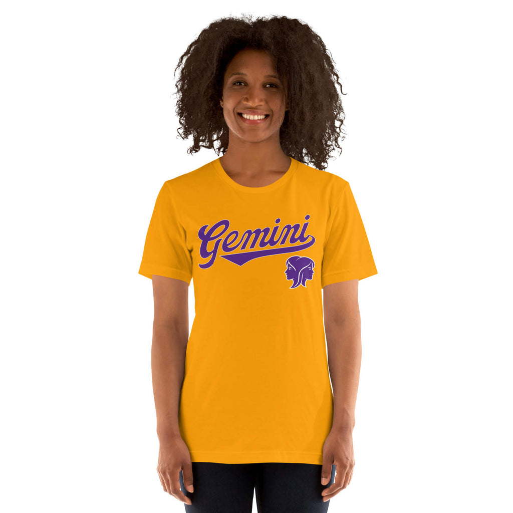 A smiling woman with curly dark hair wearing a bright gold short-sleeve t-shirt with a purple 'Gemini' script and zodiac symbol on the chest, set against a plain white background.