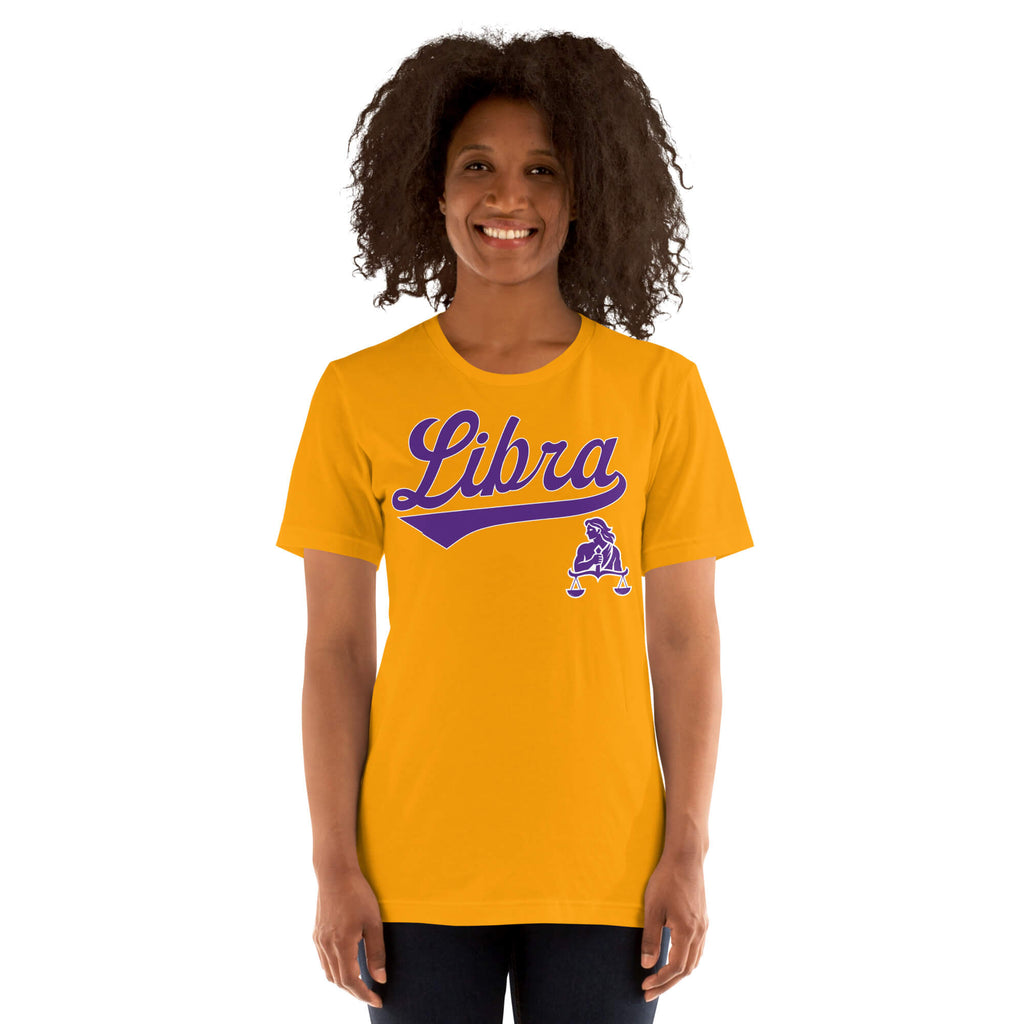 A smiling woman with curly dark hair wearing a bright gold t-shirt with a purple 'Libra' script and zodiac graphic, set against a plain white background.