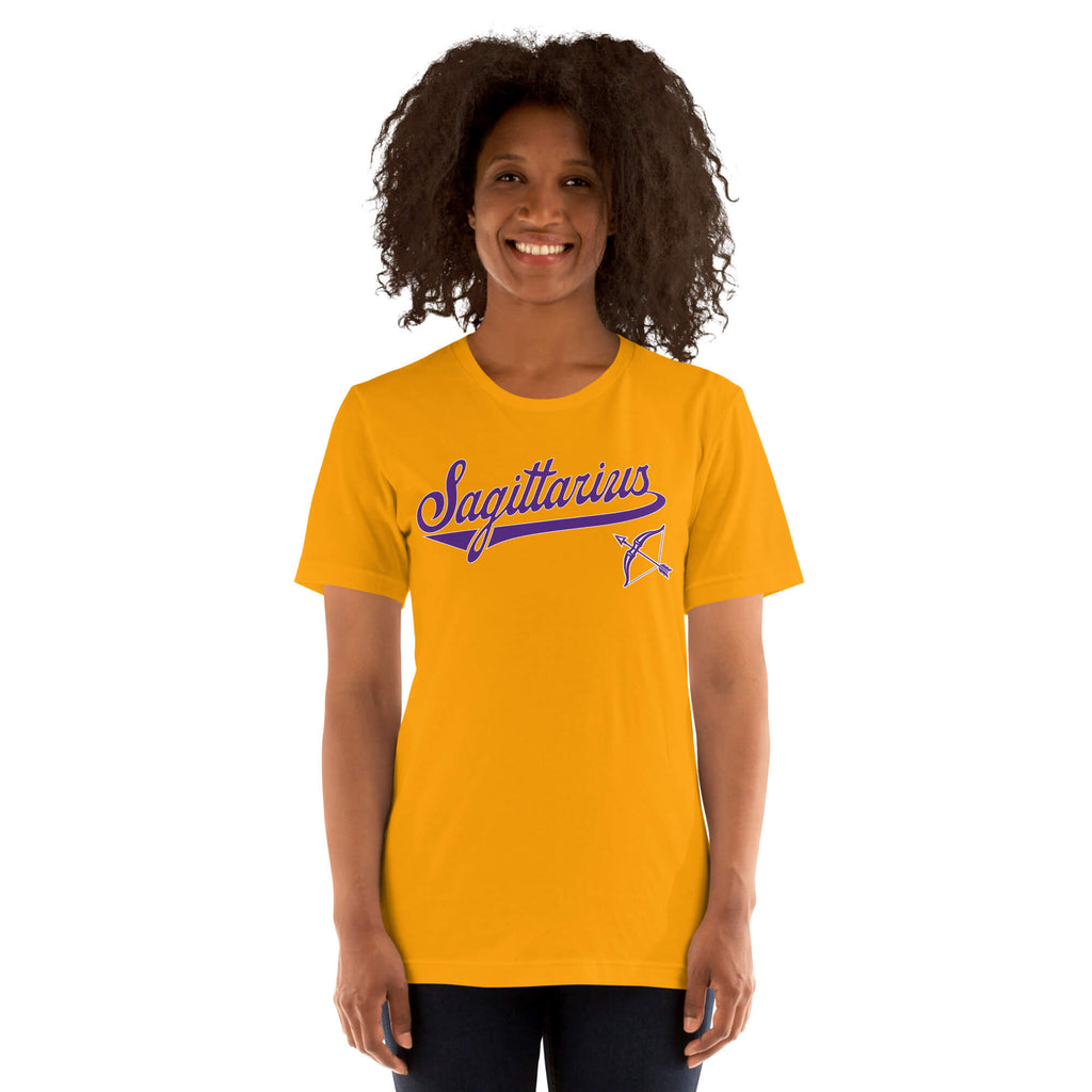 A smiling woman with curly dark hair wearing a bright gold t-shirt with a blue and purple 'Sagittarius' graphic on the chest, set against a plain white background.