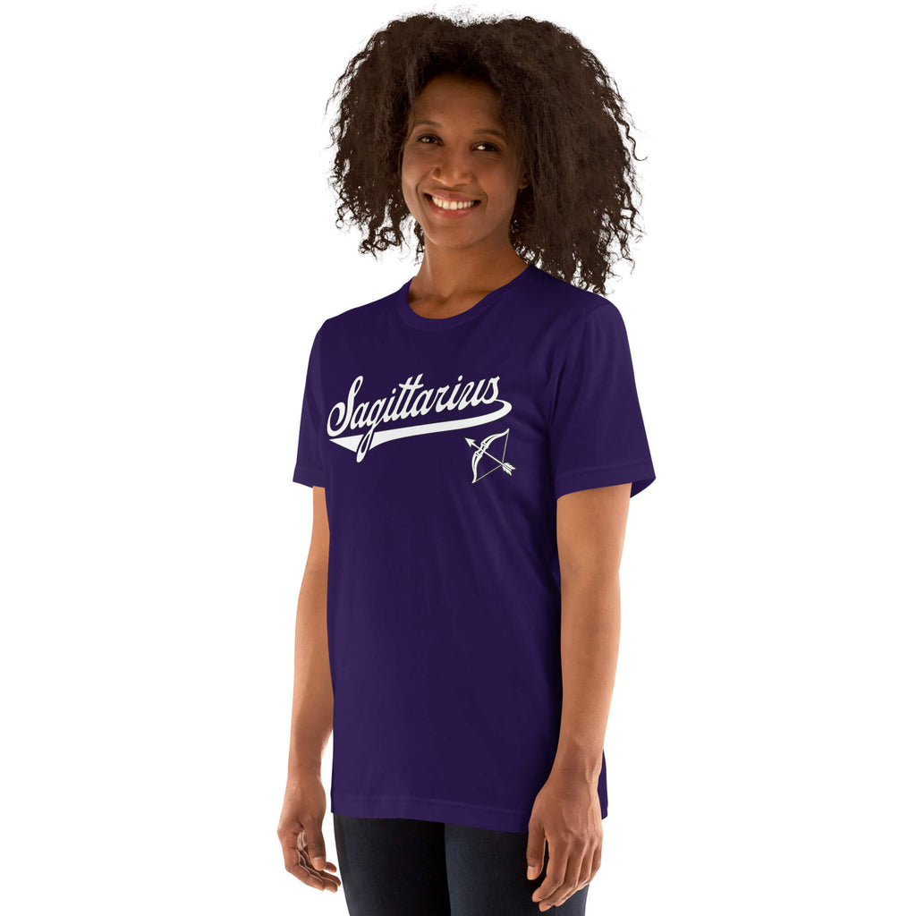 A woman with curly dark hair wearing a purple short-sleeve t-shirt with a white 'Sagittarius' script and bow graphic on the chest, shown against a plain white background.