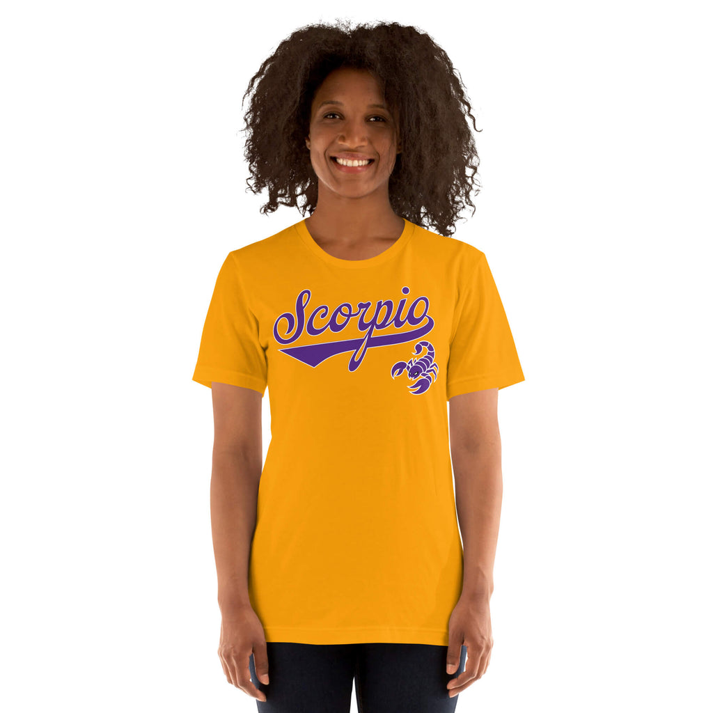 A smiling woman with curly dark hair wearing a bright gold t-shirt with a purple 'Scorpio' and scorpion graphic on the chest, set against a plain white background.