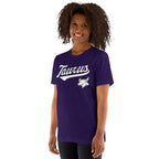 A woman with curly dark hair wearing a purple short-sleeve t-shirt with 'Taurus' in white script and a white Taurus zodiac symbol on the chest, set against a plain white background.
